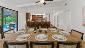 A dining area with a wooden table set for six, a ceiling fan, and a view of a kitchen and outdoor seating area. - Palm Haus • Heated Pool • Near Beach • Game Room • Volleyball • Gas Fire Pit (Palm Harbor)
