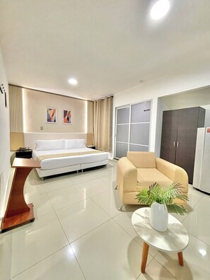 A modern living room with a white sofa, beige armchair, and a small white table with a plant. There is a large wardrobe and a bed in the background.