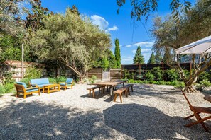 Zona salotto all'aperto con tavolo e panche, circondata da alberi e terreno sterrato.
