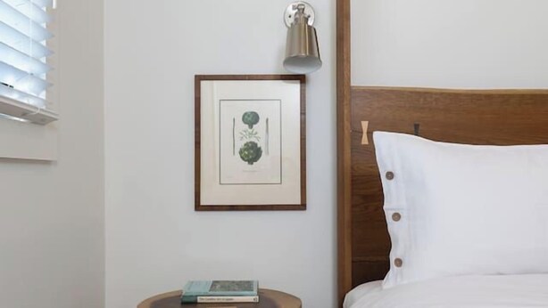 A bedroom with a wooden headboard, a bedside table with a lamp, a framed botanical print, and a small round table with books. - Jasmine residence, located downtown between the Union Hotel & Bells restaurant. (Los Alamos)