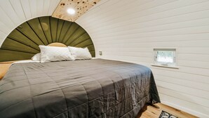 Et soveværelse med buet loft, seng med mørkt dynebetræk, hvide puder og et lille vindue.
