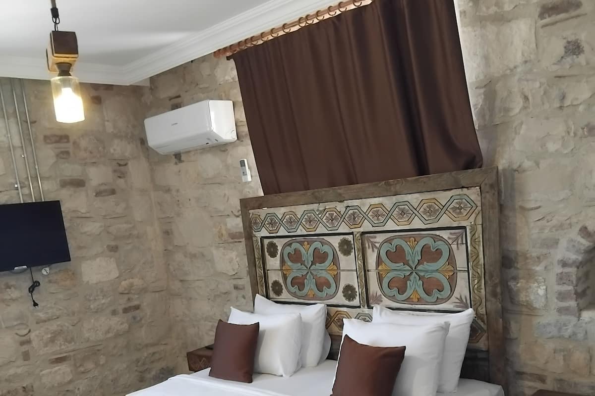 A bedroom with a stone wall, a bed with white and brown bedding, a wooden headboard with decorative tiles, a brown curtain, a mounted TV, and a yellow chair.