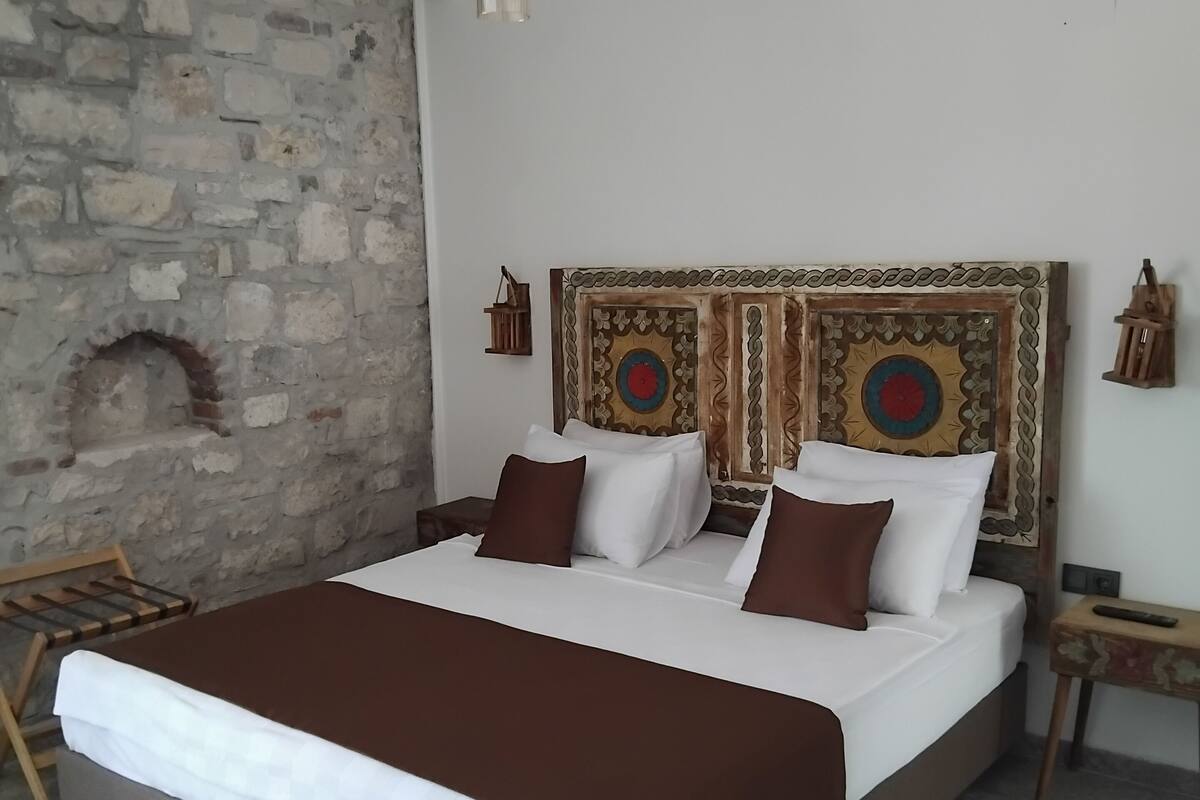 A bedroom with a stone wall, a bed with white and brown bedding, a red and white rug, and a wooden headboard with decorative patterns.
