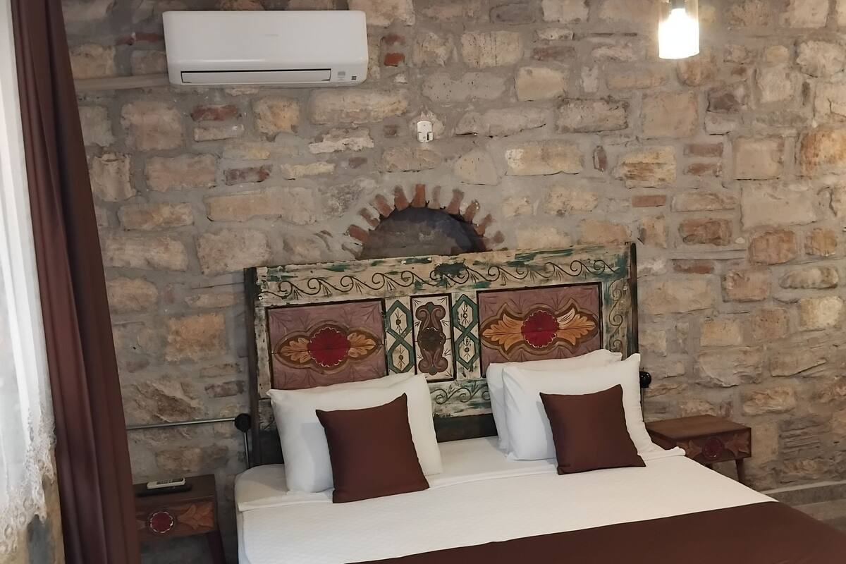 A bedroom with a stone wall, a decorative headboard, a bed with white and brown bedding, and a ceiling light fixture.
