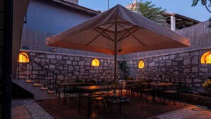 Een terras buiten met stenen muren, houten vloeren en tafels en stoelen onder een grote parasol.