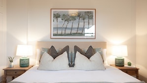 A hotel room with a bed, two bedside tables with lamps, a framed picture of palm trees, and a small plant on one table.