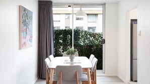 A modern dining area with a white table and chairs, a hanging plant, and a view of a building through a large window.