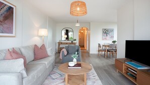 A modern living room with a grey sofa, a round wooden coffee table, and a flat-screen TV. There is a dining area with a table and chairs, and a hallway leading to another room.