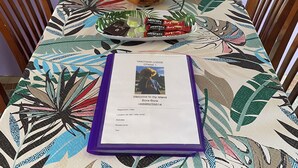 Een tafel met een tafelkleed met een tropisch bladpatroon, een fruitschaal, een vaas met bloemen en een welkomstfolder.