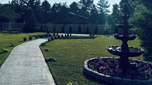 A pathway leading to a building with a fence, surrounded by trees and a fountain.