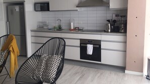 Eine moderne Küche mit weißen Schränken, einem Edelstahlkühlschrank, einer Mikrowelle und einem Einbauofen. Zwei schwarze Stühle mit gemusterten Kissen und eine gelbe Decke stehen dort.