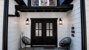 A black door with frosted glass panels, flanked by two black metal chairs and two wall-mounted lanterns. A welcome sign is positioned to the left of the door. - Beautiful, Spacious 4-BR  home in Thackerville, OK near Winstar World Casino (Thackerville)