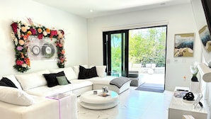 A modern living room with a white sofa, a coffee table, and a large window with a view of greenery. - 5bdr/5.5ba Modern Oasis with outdoor Gym, Cold Plunge, Heated Pool & Jacuzzi (Naples)