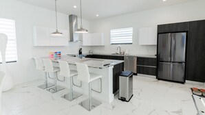 A modern kitchen with a central island, white bar stools, stainless steel appliances, and a marble backsplash. - 5bdr/5.5ba Modern Oasis with outdoor Gym, Cold Plunge, Heated Pool & Jacuzzi (Naples)