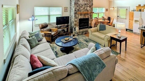 A spacious living room with a stone fireplace, a ceiling fan, and a view of the kitchen. - Charming and Cozy River House (Murphy)