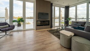 A spacious living room with a fireplace, large windows, and a view of a lake. - Luxury Villa With Dishwasher (Kerkdriel)