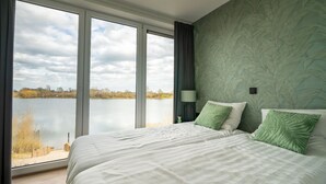 A bedroom with a large window overlooking a lake, a bed with white bedding, and a green leaf-patterned wall. - Luxury Villa With Dishwasher (Kerkdriel)
