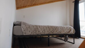 Ein Einzelbett mit einem gemusterten Bettbezug in einem Zimmer mit Holzbalkendecke und einem Fenster mit Vorhängen.
