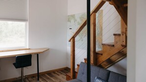 Ein Zimmer mit einem Holztisch, einem schwarzen Stuhl, einem grauen Sofa und einer Holztreppe. An der Wand hängt eine Karte.