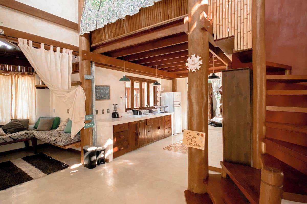 A spacious living area with a high ceiling, wooden beams, a staircase, and a kitchen area.
