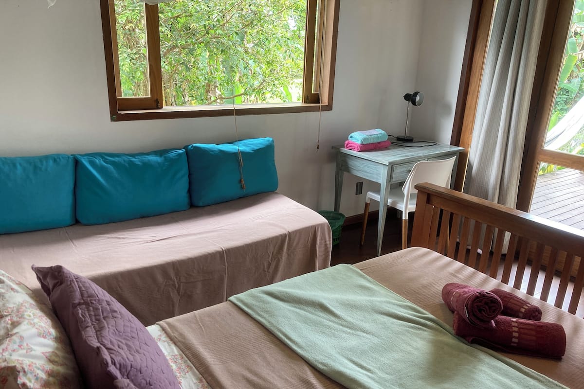 A room with a bed, a desk, and a chair. There is a window with a view of greenery outside.