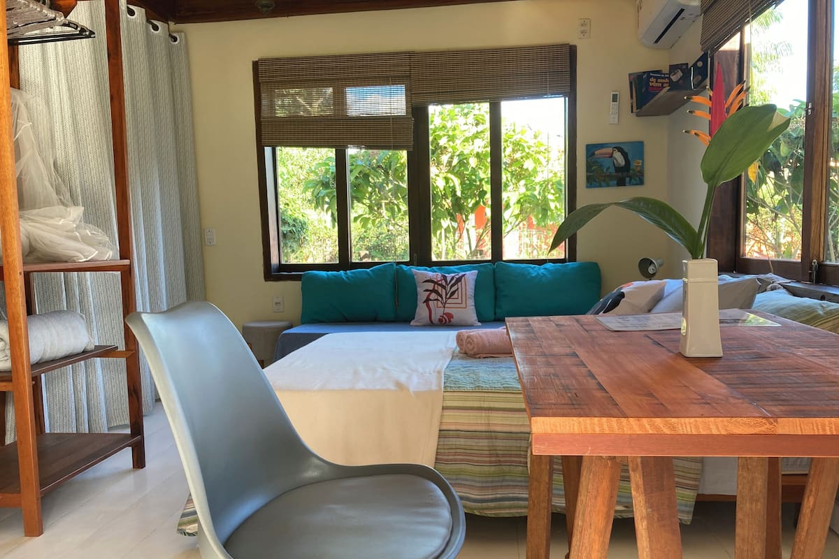 A cozy room with a bed, a wooden table, a chair, and a window with a view of greenery.