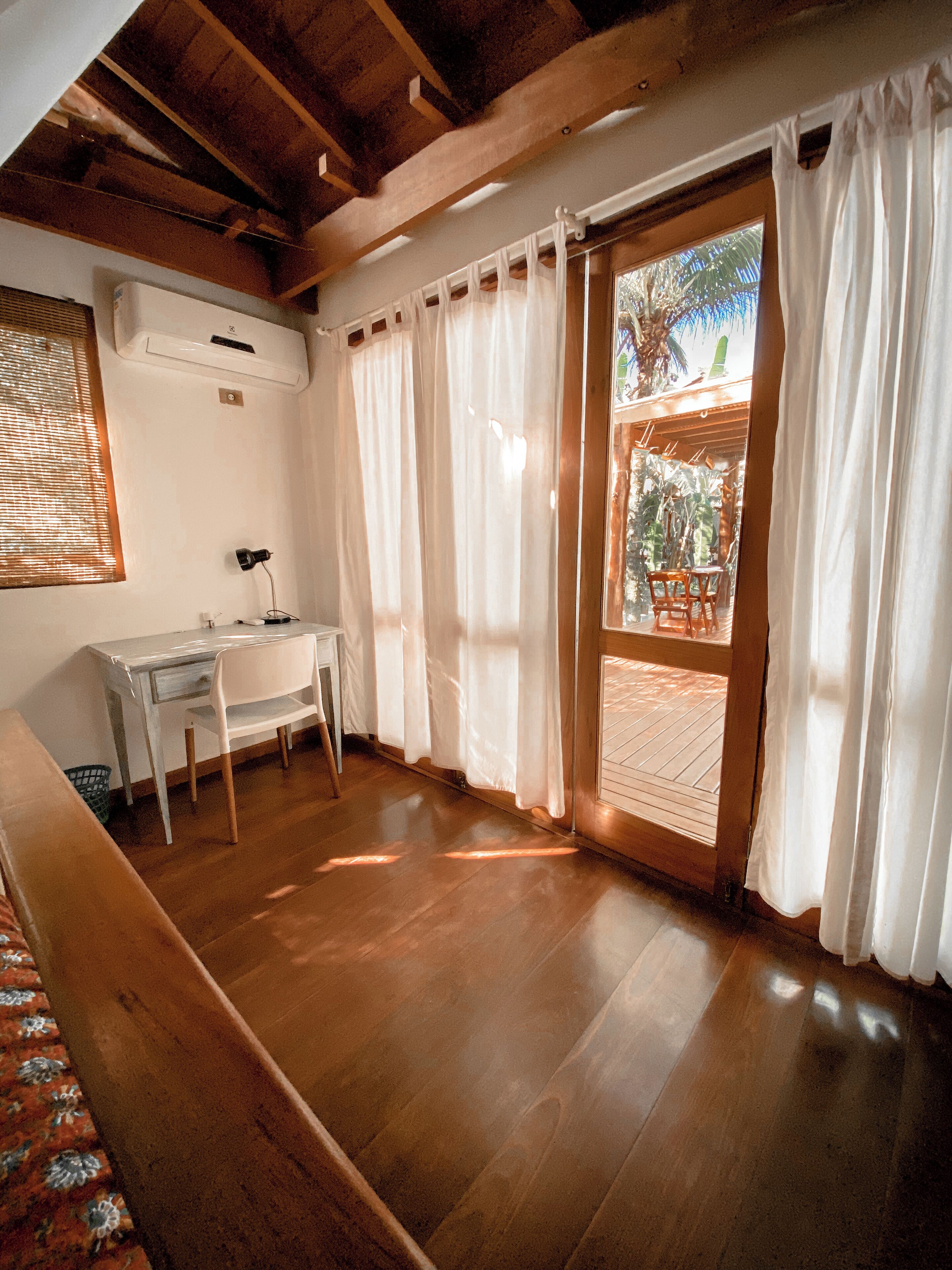 A room with wooden floors, a desk, and a chair. There is a window with blinds and a door leading outside.