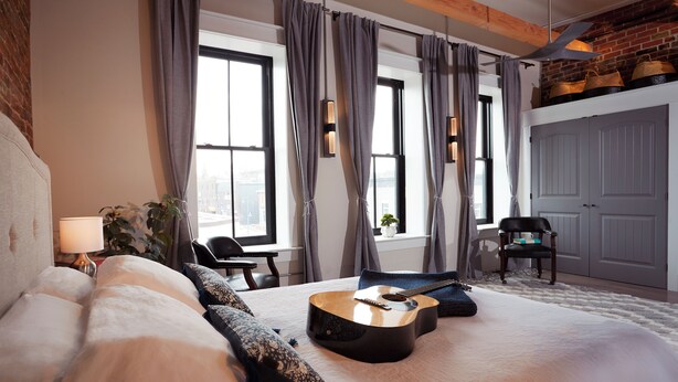 A bedroom with a bed, a guitar, a chair, a desk, and a view of the city through large windows. - French Inn in Downtown Belleville ,  right above our charming French Bistro  (Belleville)