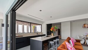 Una cocina moderna con una isla central, armarios negros y un ventanal grande. Hay un área de comedor con un sofá marrón y taburetes blancos.