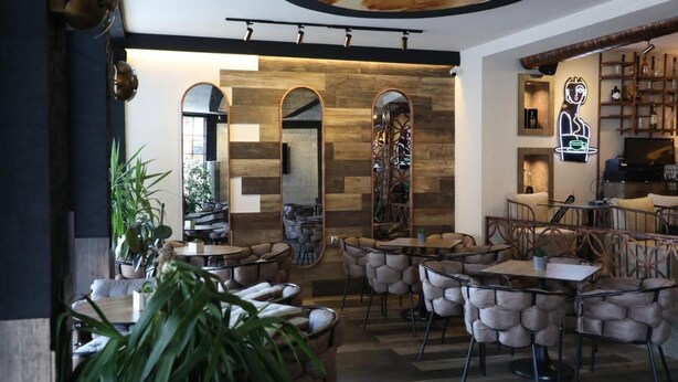 A modern restaurant interior with round tables and cushioned chairs, a wooden accent wall, and a neon sign. - Bruti Hotel (Prizren)