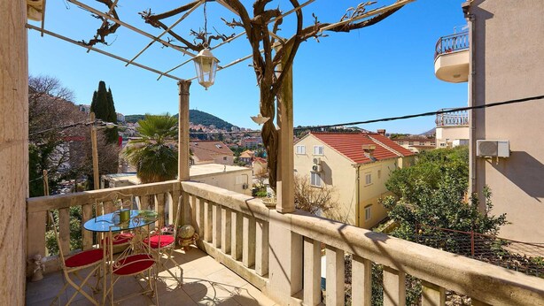 Un balcon avec une table et des chaises, un arbre, et une vue sur des bâtiments et une colline en arrière-plan.