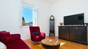 Un salon avec un canapé rouge, une table basse en bois, un meuble noir sur lequel se trouve un téléviseur, et une grande fenêtre donnant sur des collines.