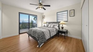 A bedroom with a bed, two bedside tables, a ceiling fan, a window with blinds, and a balcony door.