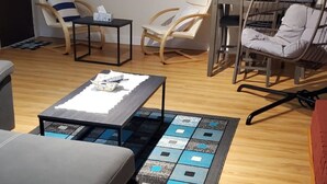 A modern living room with a grey sofa, a coffee table, and a rug with a geometric pattern. There are two rocking chairs and a small table with a laptop. Large windows offer a view of the sunset.