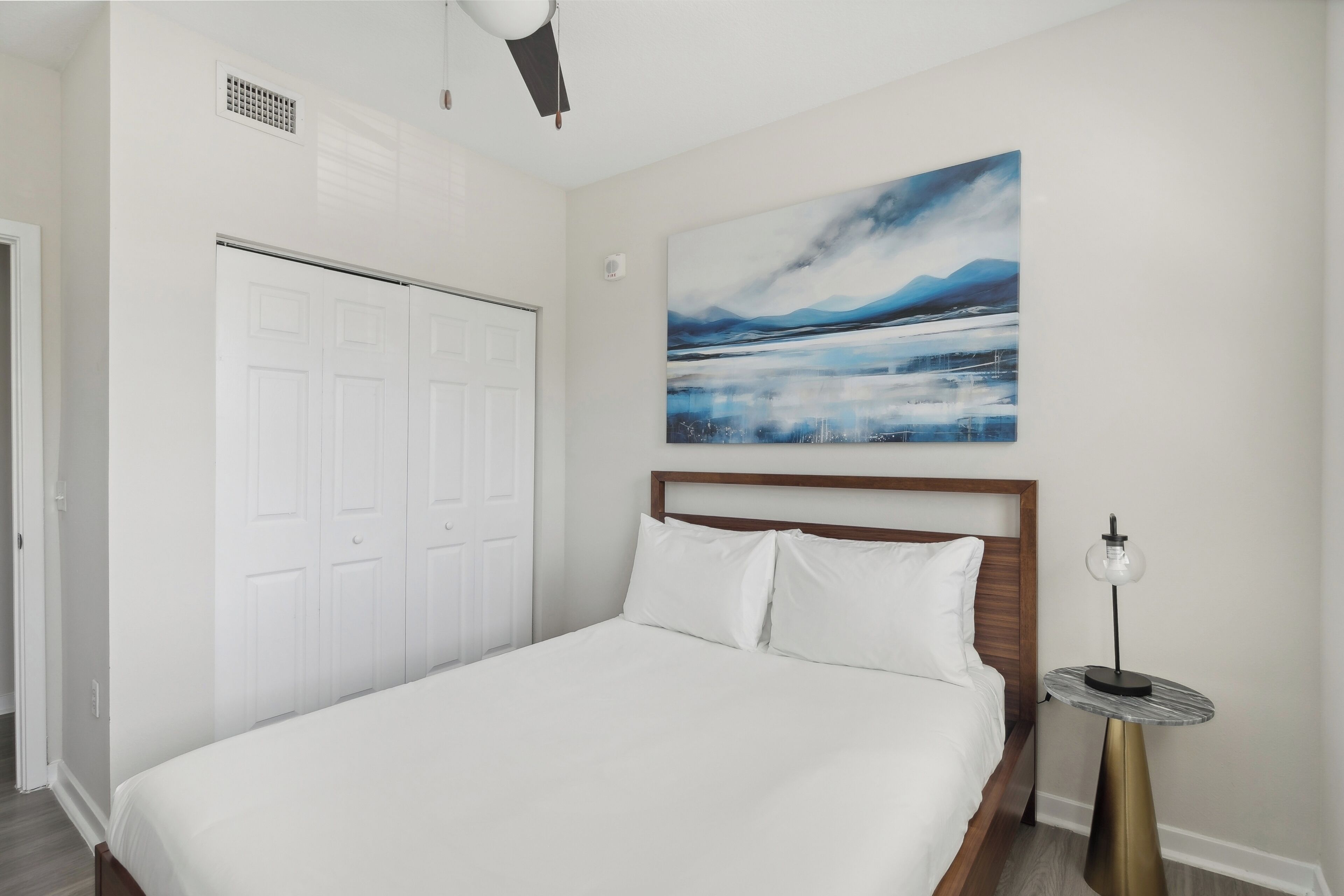 A bedroom with a bed, a white wardrobe, a painting, and a bedside table with a lamp.