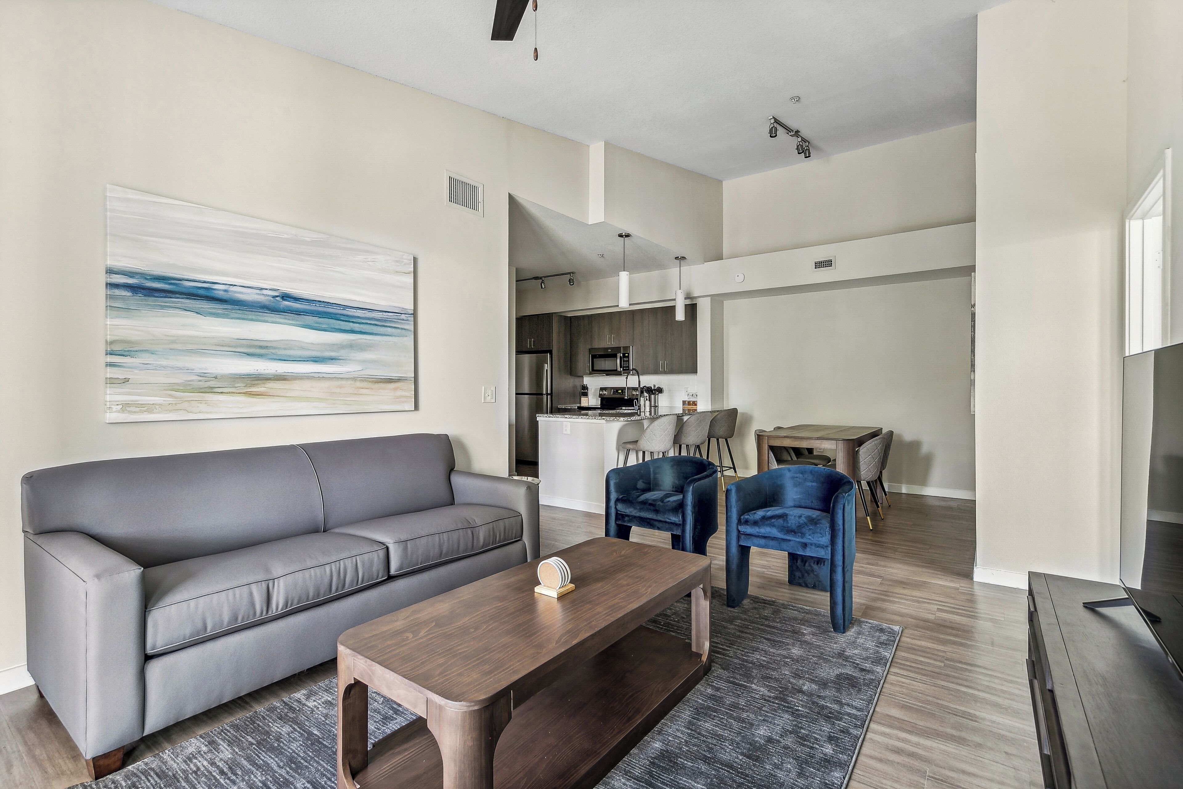 Una sala de estar moderna con un sofá gris, una mesa de centro de madera y una silla azul. Se observa una cocina al fondo con una isla blanca y un comedor con mesa y sillas.