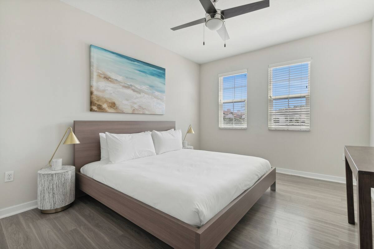 A bedroom with a bed, a ceiling fan, a painting, and two windows.
