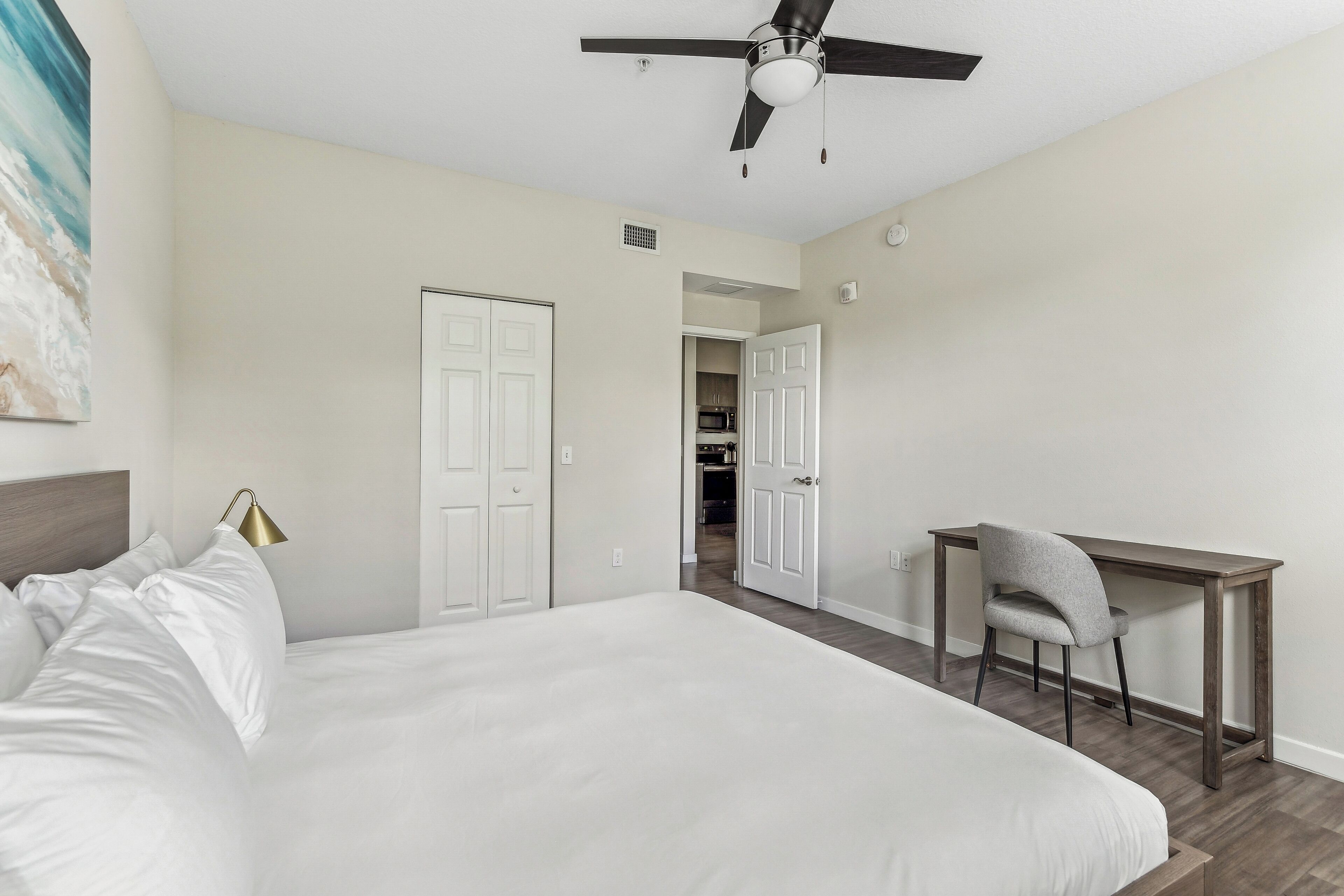A bedroom with a bed, a desk, and a chair. There is a ceiling fan, a door, and a wall-mounted lamp.