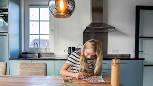 Uma cozinha moderna com uma mulher desenhando numa mesa de madeira, um lustre e uma janela com vista.