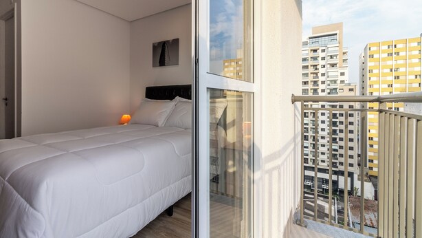 A bedroom with a bed, a bedside table, and a window with a view of buildings. - Akile Bela Vista Tatro Renault (São Paulo)