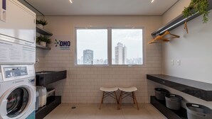 A modern laundry room with a washing machine, two white chairs, a window with a city view, and a shelf with plants and hangers. - Akile Bela Vista Tatro Renault (São Paulo)