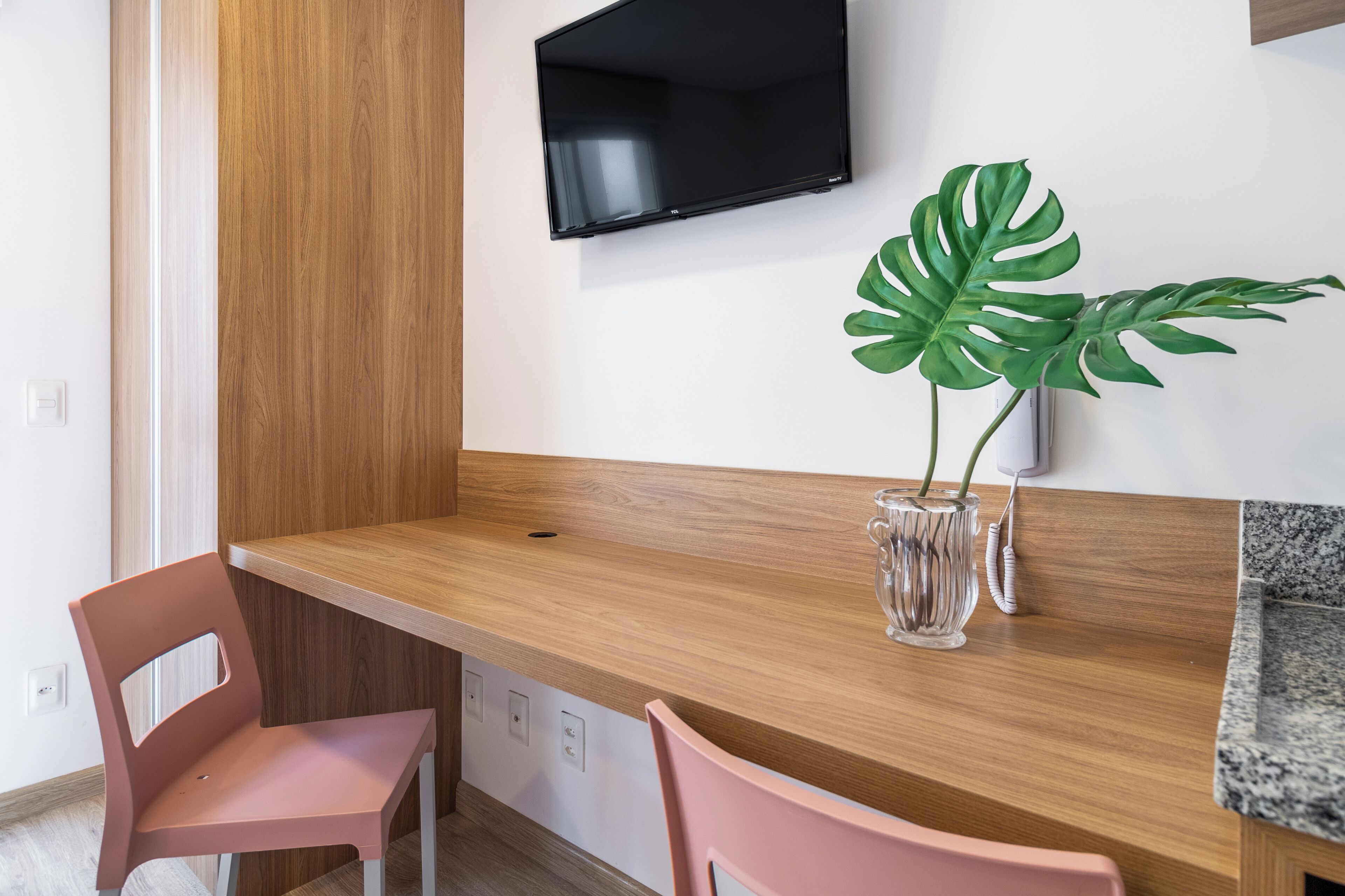 Una habitación moderna con un escritorio de madera, dos sillas rosas, una planta verde grande y un televisor de pantalla plana montado en la pared.