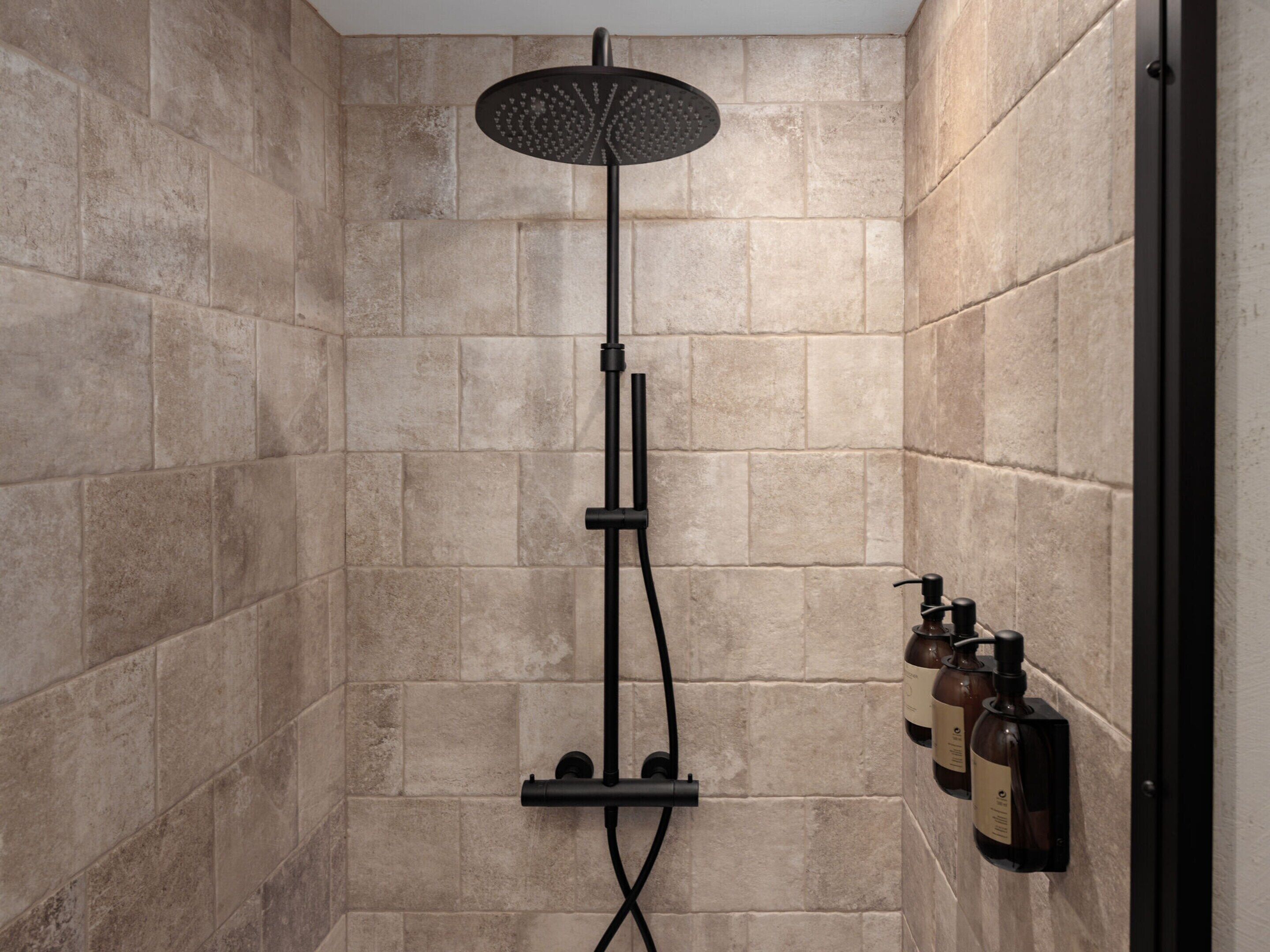 A shower with a rain showerhead, a handheld showerhead, and a wall-mounted control panel with bottles.