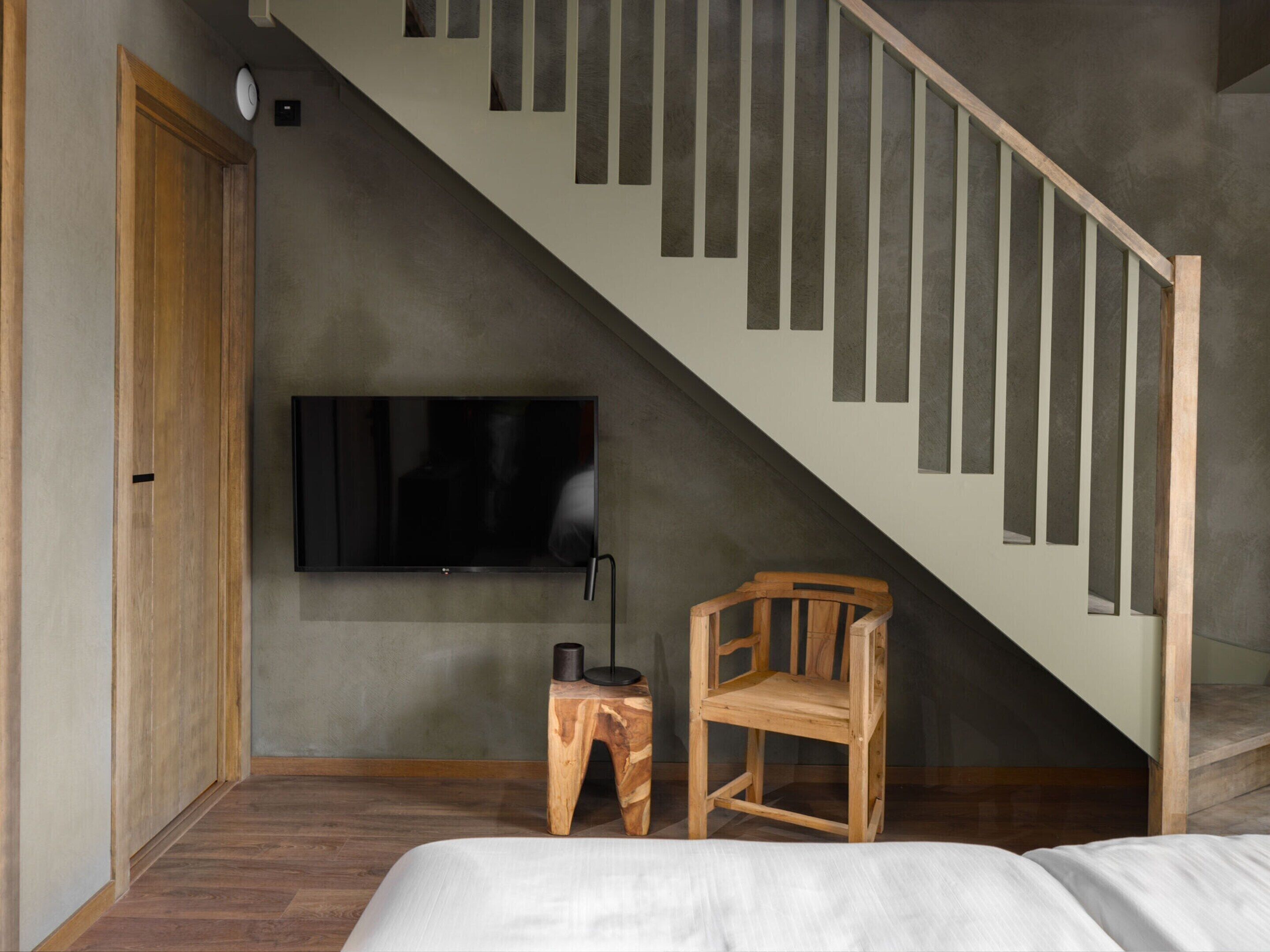 Un dormitorio con una silla de madera, una mesita auxiliar, una cama, un televisor montado en la pared y una escalera con barandillas de madera.
