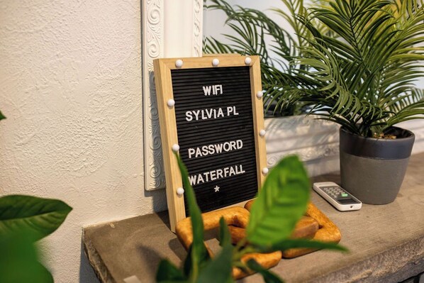 A sign with "WIFI SYLVIA PL PASSWORD WATERFALL" on a wooden board with white dots, placed on a stone ledge with a potted plant and a remote control.
