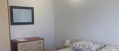 A hotel room with a bed, a bedside table, a lamp, a mirror, and a wooden dresser.