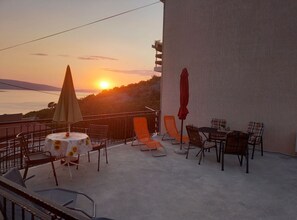 Een terras met een tafel en stoelen, een rode parasol, en uitzicht op de ondergaande zon boven de zee.