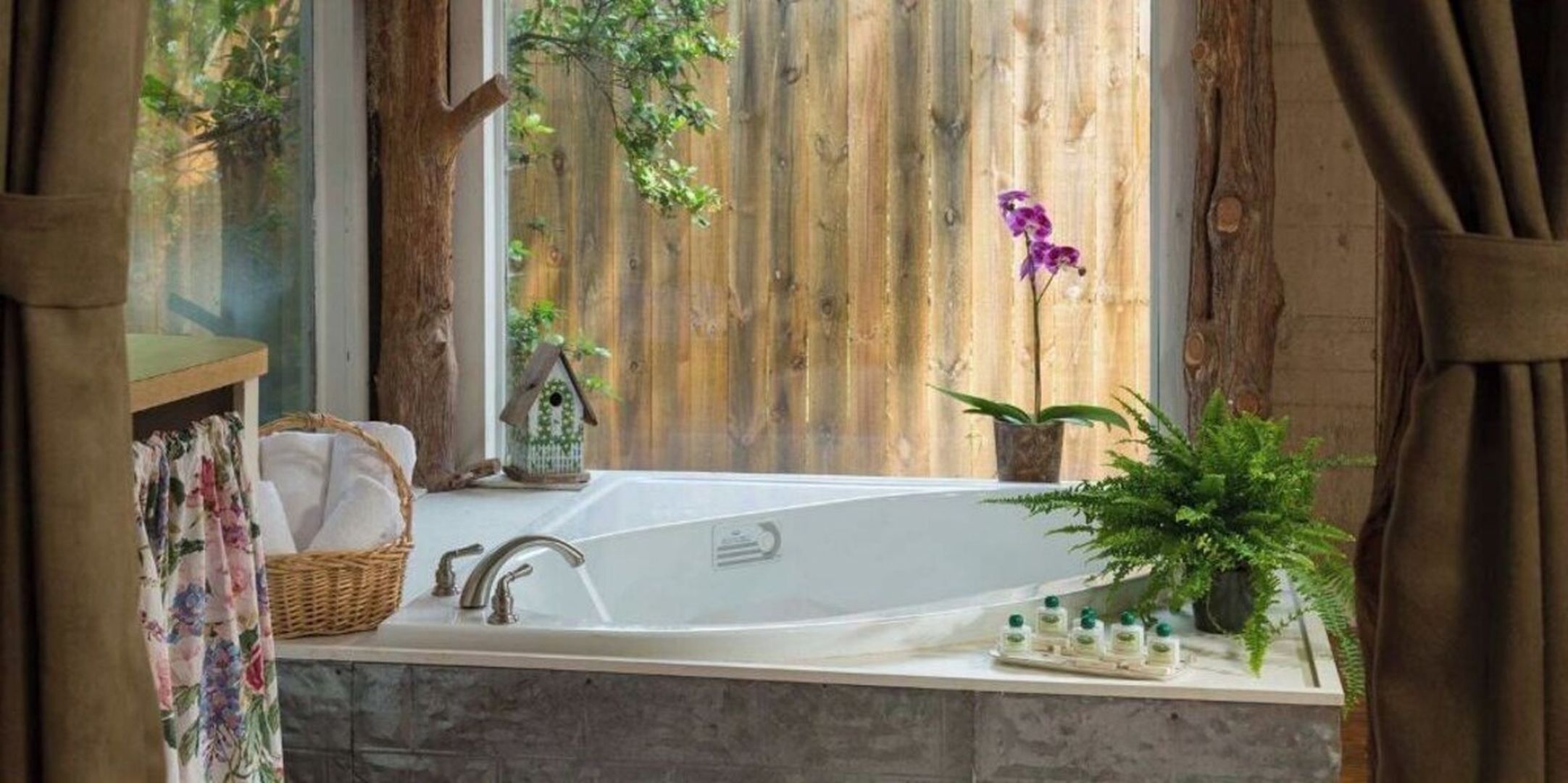 Un baño con bañera, ventana con vistas a una cerca de madera y vegetación, una cesta de mimbre con toallas y una pequeña planta en maceta.