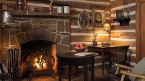 A cozy cabin interior with a stone fireplace, wooden walls, a table with a bowl of fruit, and a chair. - 1800-style cedar Log Cabin on Blisswood Ranch with many optional activities! (Cat Spring)