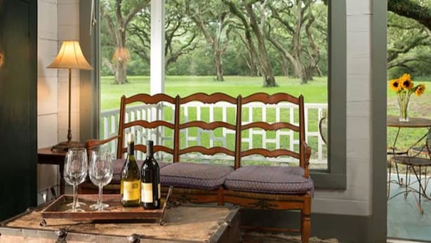Un banc en bois, avec en arrière-plan une clôture blanche en bois, une table ornée de bouteilles de vin et de verres, et une vue sur un parc verdoyant et luxuriant, visible à travers de grandes fenêtres.